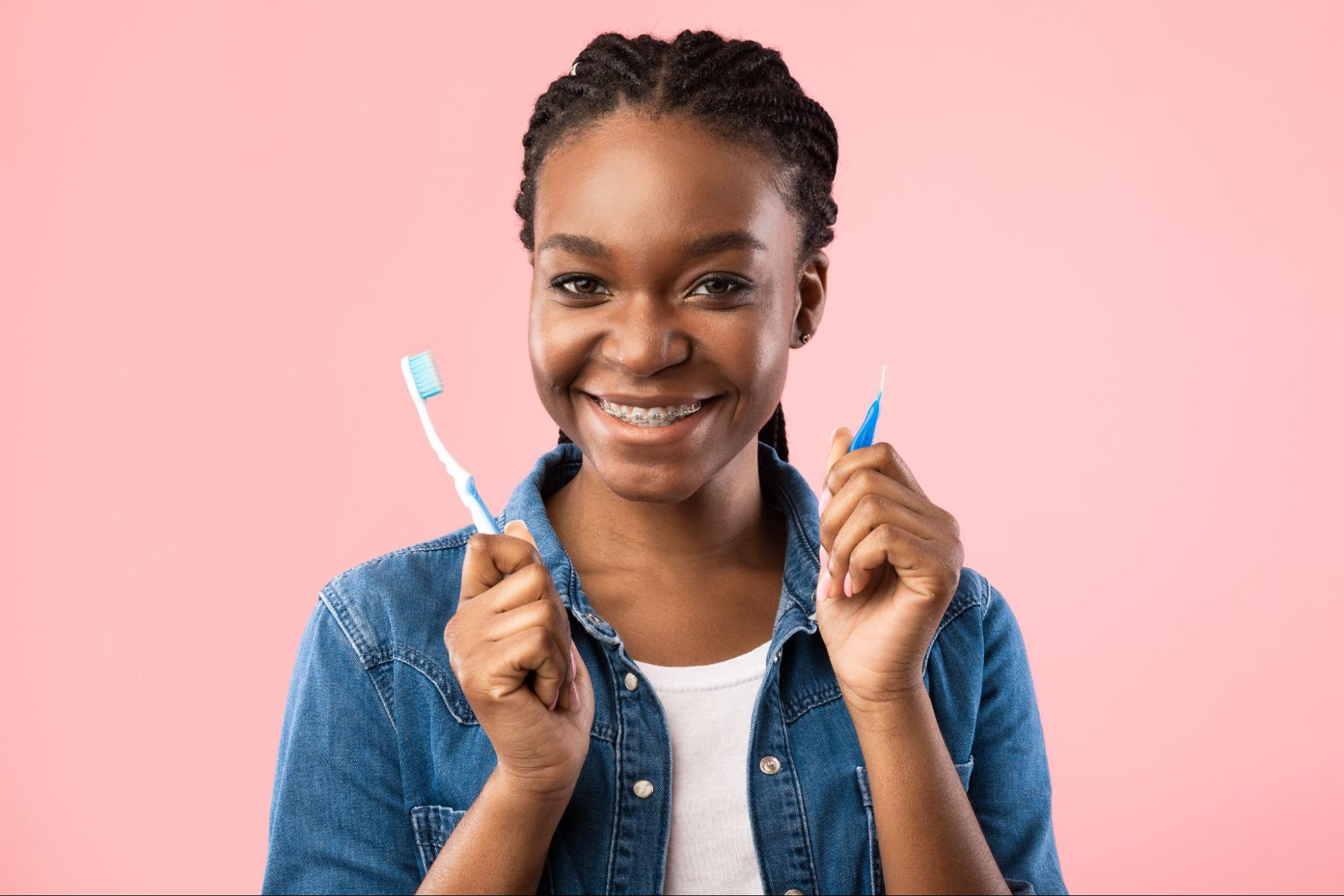 Smiling girl with braces holding a toothbrush and an interdental brush, promoting oral hygiene for braces and clear aligners at Viva Orthodontics in Bountiful.