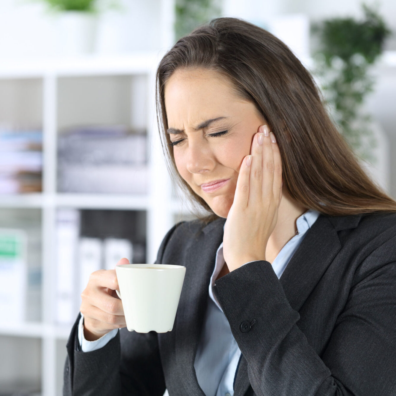 Woman in business attire holding a cup, experiencing jaw pain and discomfort, representing TMJ issues related to orthodontic care at Viva Orthodontics in Bountiful.